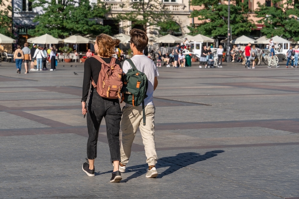 Młoda para z plecakami spacerująca po rynku, trzymając się za ręce.